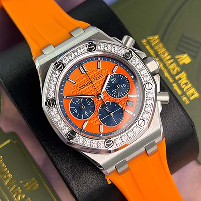 Reloj Audemars Piguet Royal Oak con esfera naranja, subesferas azules, correa de caucho y bisel con brillantes, estilo deportivo y moderno.