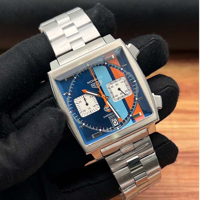 Reloj TAG Heuer Monaco Gulf Racing Classic con esfera azul y franjas naranja y celeste, caja cuadrada y correa metálica.