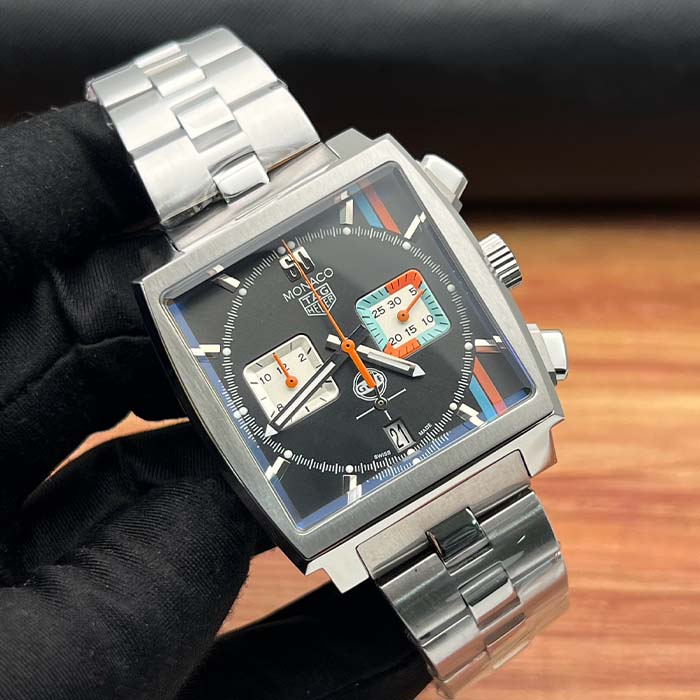 Reloj TAG Heuer Monaco con carátula negra, detalles en naranja y celeste, diseño cuadrado deportivo y correa metálica.